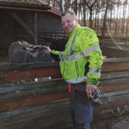 Krzysztof Roguski - Uśmiechnięty mężczyzna w odblaskowej kurtce głaszcze osła za drewnianym ogrodzeniem. W tle budynek gospodarczy i drzewa.