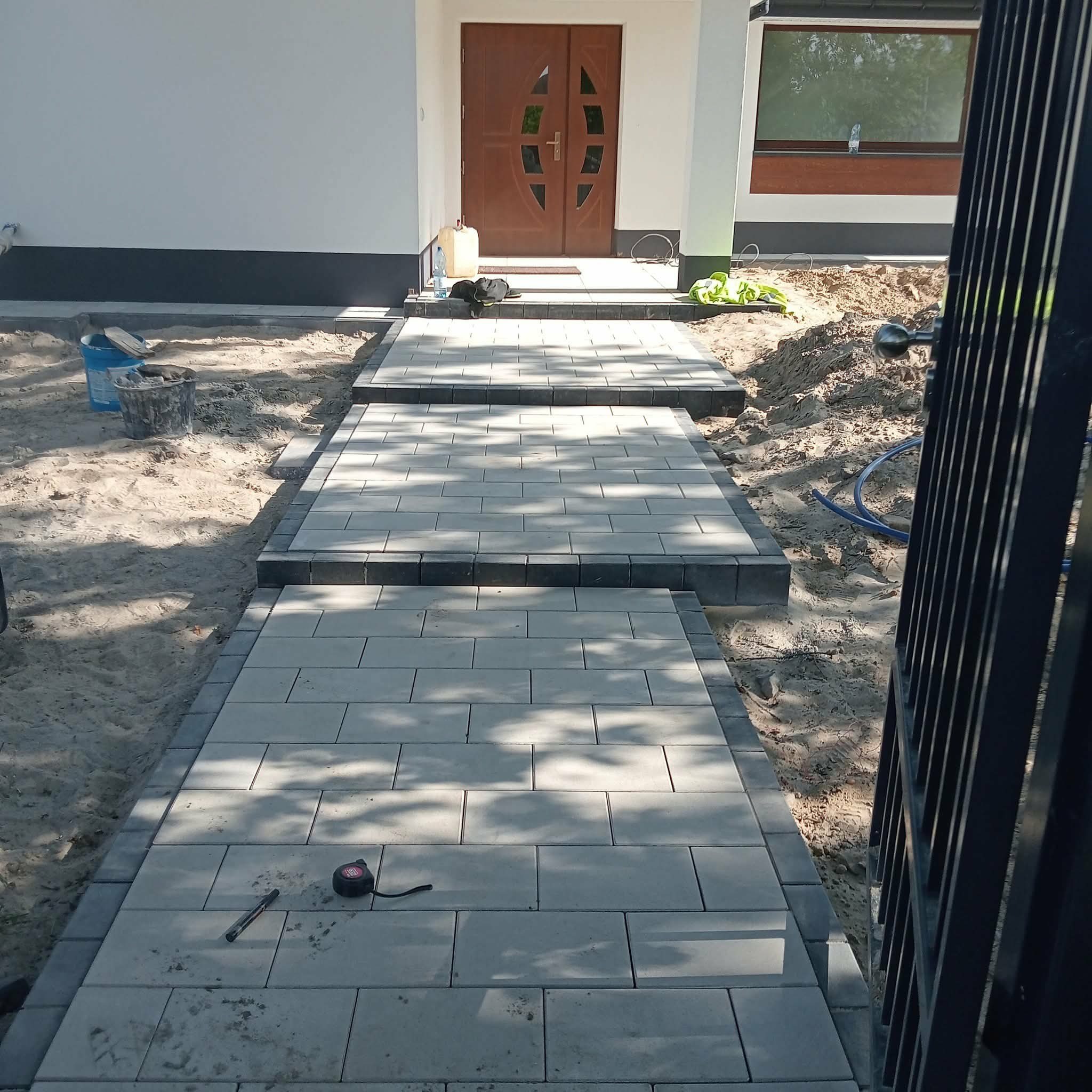 Ścieżka z jasnoszarej kostki brukowej prowadząca do drzwi wejściowych domu, obrzeża z ciemniejszej kostki. Widoczne narzędzia pracy i fragment ogrodzenia.