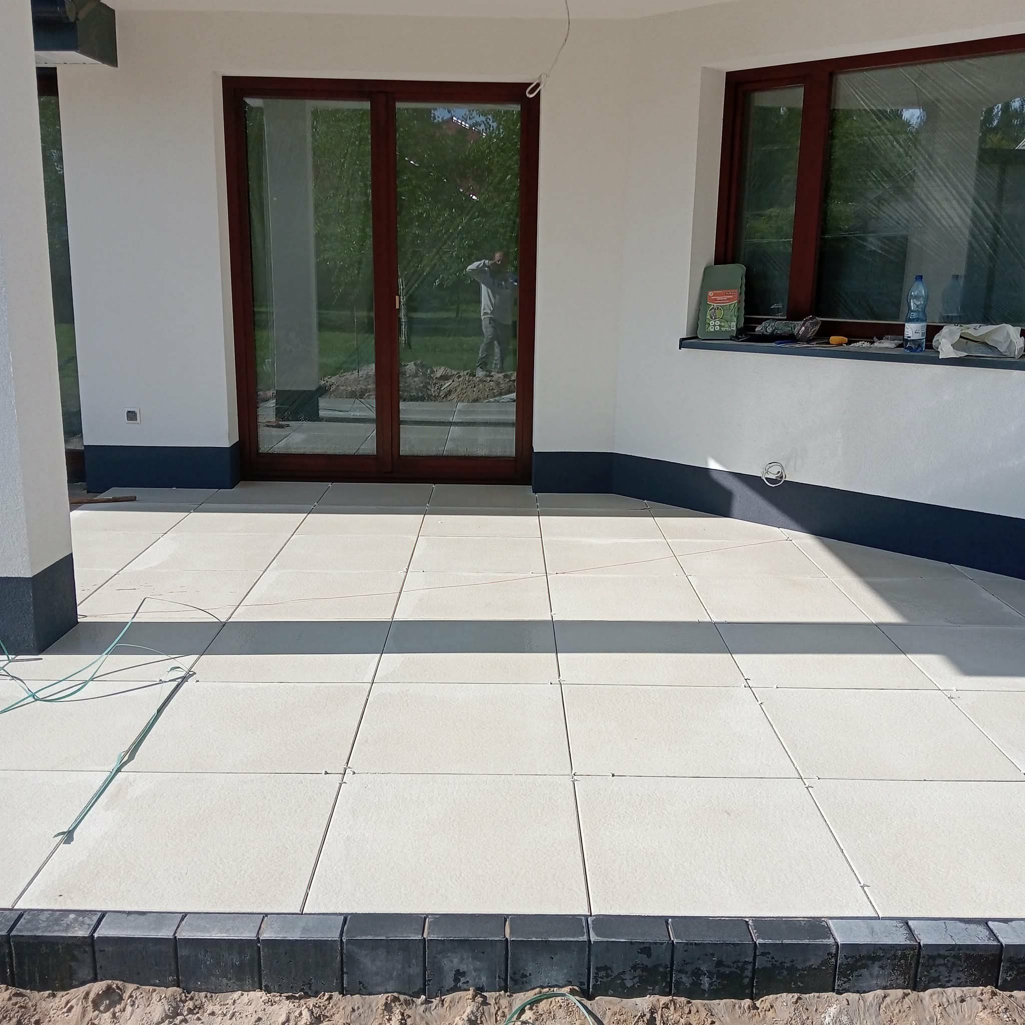 Jasne płyty tarasowe ułożone na zewnątrz budynku z ciemną podmurówką i oknami. Widoczne fugi i obrzeża z kostki. W tle odbicie osoby w oknie.