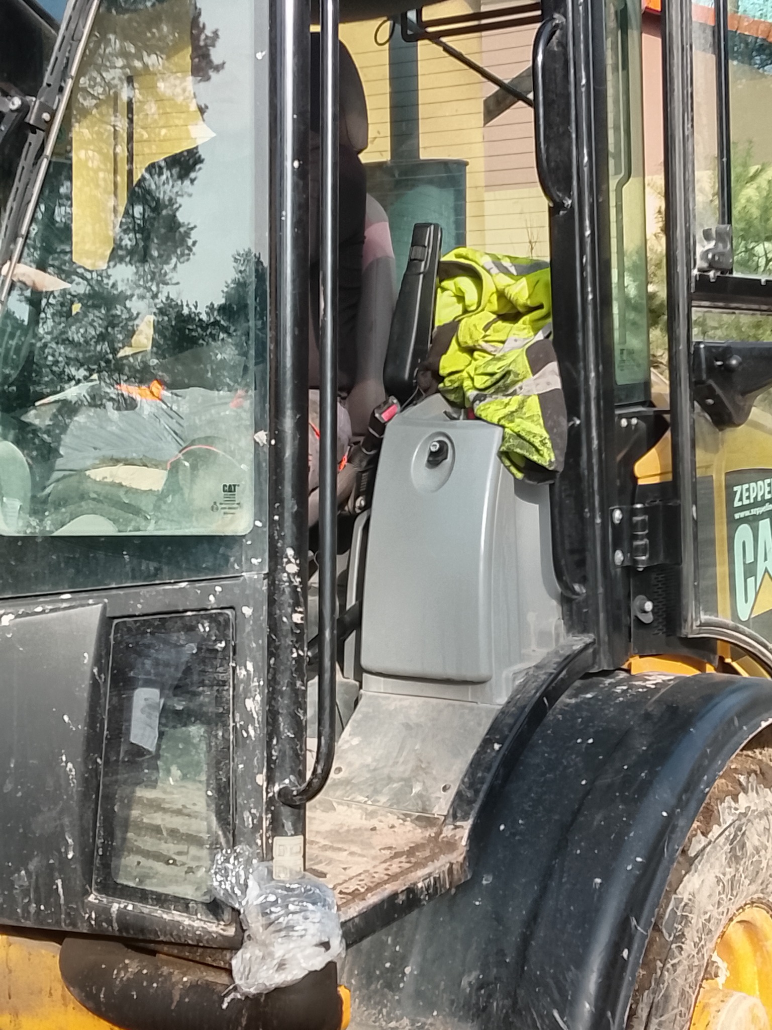 Wnętrze kabiny brudnej koparko-ładowarki marki CAT, widoczne elementy sterowania, siedzenie i odblaskowa kamizelka. Na szybie naklejka z logo CAT.