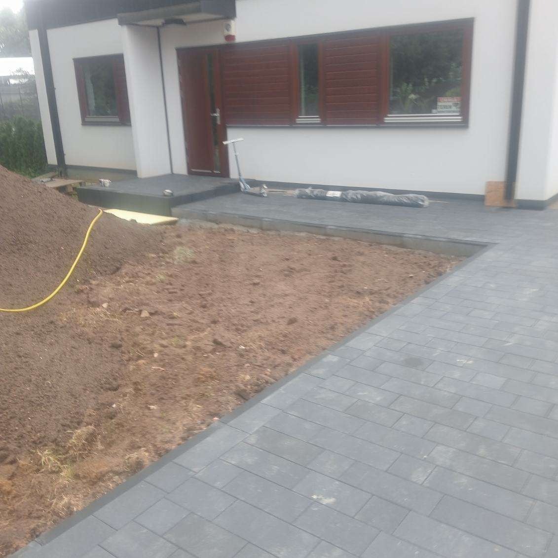 Częściowo ułożona, szara kostka brukowa przed domem z brązowymi okiennicami. Widoczna sterta ziemi i przygotowany teren pod dalsze prace brukarskie.