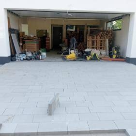 Jasnoszara kostka brukowa na podjeździe garażowym, z widocznymi narzędziami i materiałami budowlanymi w tle. Ujęcie z niskiego kąta, podkreślające wzór kostki.