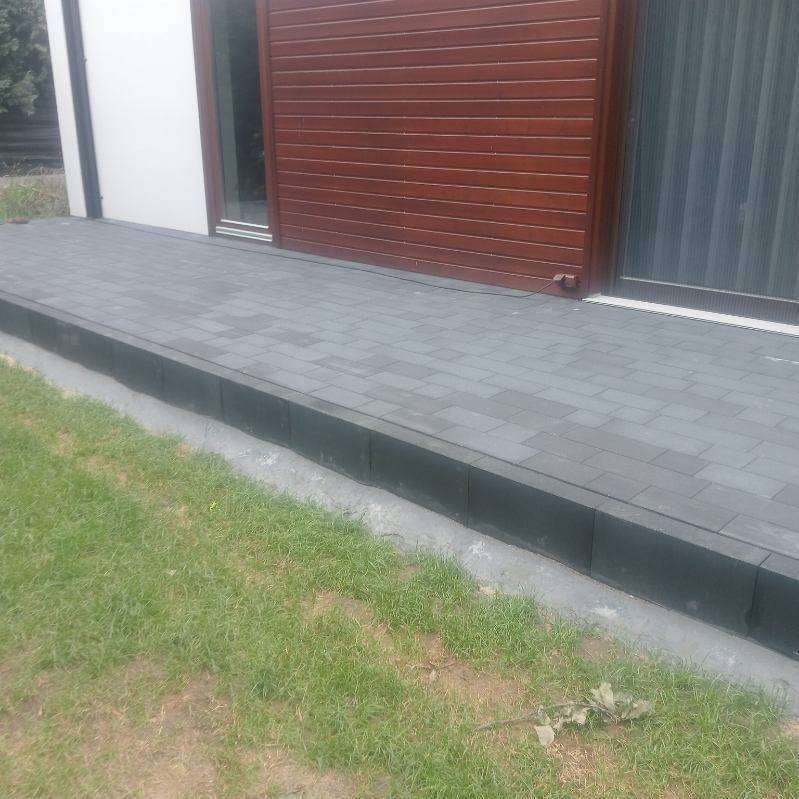 Szara kostka brukowa ułożona przy budynku z drewnianą elewacją i trawnikiem. Widoczne obrzeża z ciemnej kostki. Ujęcie z poziomu gruntu.