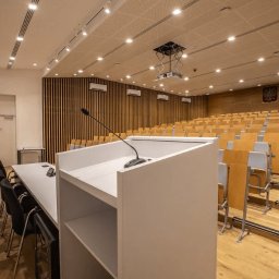 Pro Acoustic Ireneusz Tolak - Nowoczesna aula uniwersytecka z drewnianymi elementami, oświetleniem LED i białą mównicą z mikrofonem. Widoczne rzędy krzeseł i godło Polski na ścianie.