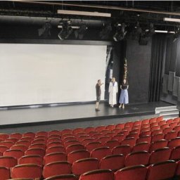 Pro Acoustic Ireneusz Tolak - Widok na scenę teatru z rzędami czerwonych foteli. Na scenie trzy osoby, oświetlenie punktowe. Ściany w ciemnych kolorach, akustyczne panele po prawej.
