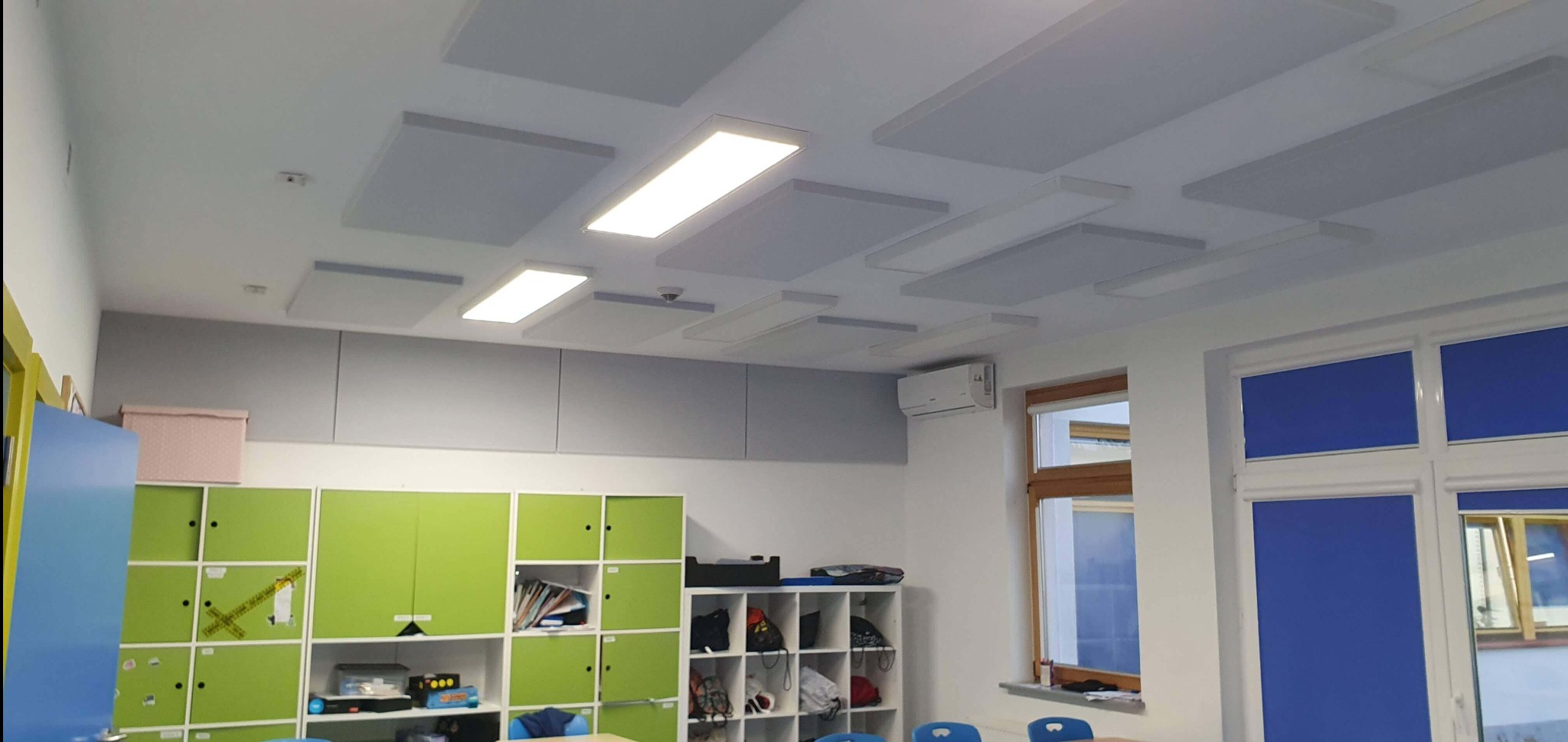 Sala lekcyjna z panelami akustycznymi na suficie i oświetleniem LED. Widoczne szafki w kolorze zielonym i szarym oraz okno z niebieską roletą.