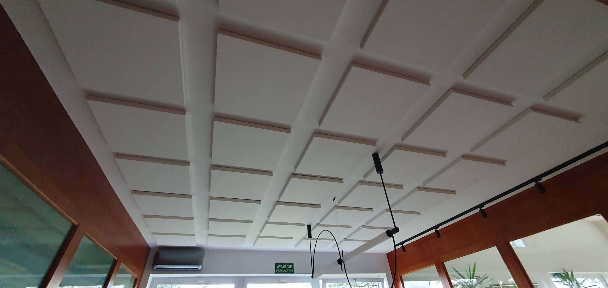 Biały sufit kasetonowy z drewnianymi listwami, widok z dołu, oświetlenie punktowe i wiszące lampy w tle. Wykończenie wnętrza biura lub lokalu usługowego.