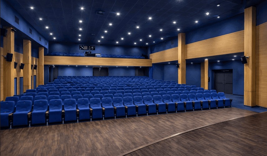 Wnętrze sali kinowej lub teatralnej z rzędami niebieskich foteli, drewnianą podłogą i panelami ściennymi. Oświetlenie punktowe na ciemnoniebieskim suficie.