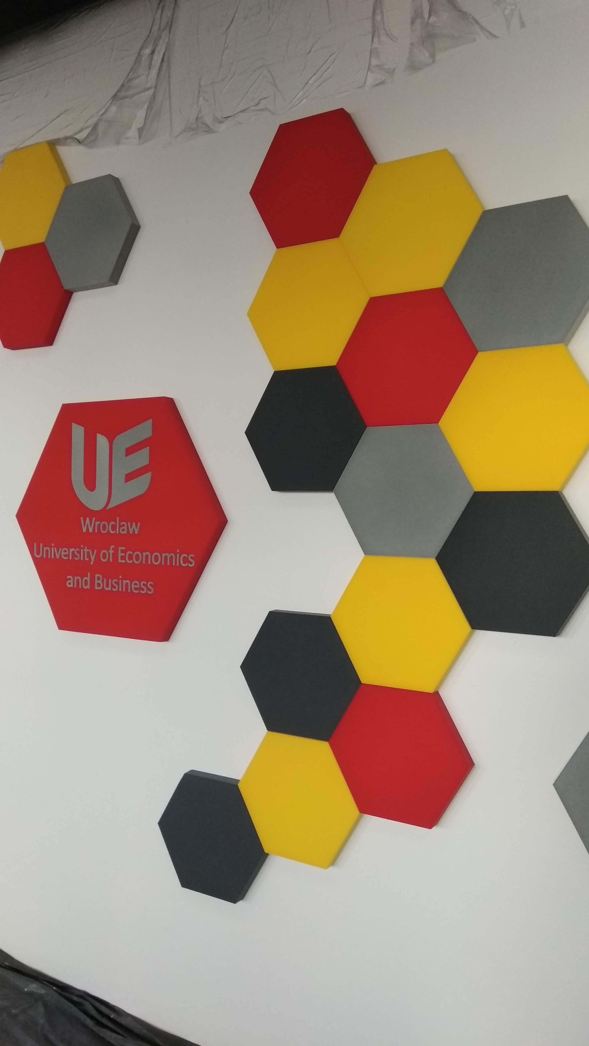 Dekoracyjne panele akustyczne w kształcie heksagonów w kolorach: czerwonym, żółtym, szarym i czarnym, zamontowane na białej ścianie w biurze Uniwersytetu Ekonomicznego we Wrocławiu.