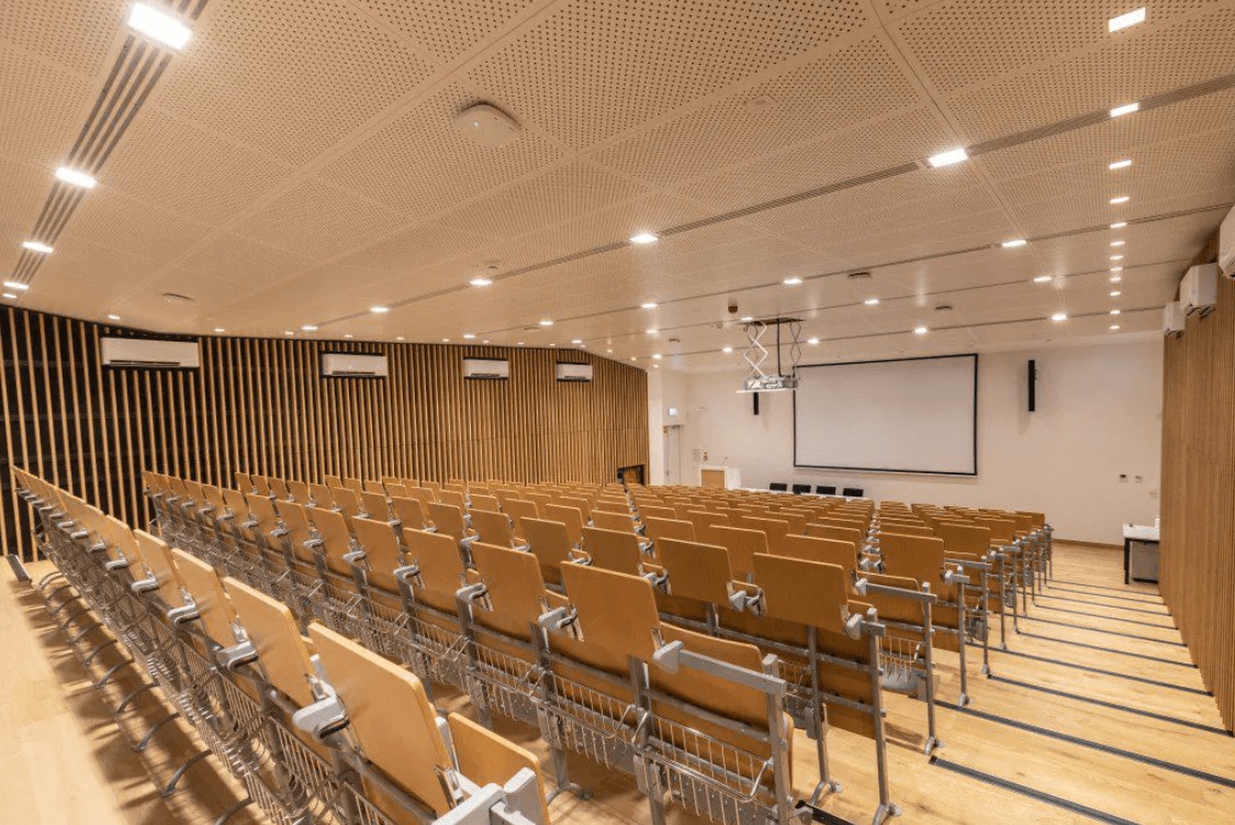 Sala konferencyjna z rzędami krzeseł, drewnianą podłogą i ścianami z paneli. Sufit podwieszany z oświetleniem. Widoczny ekran projekcyjny i klimatyzacja.