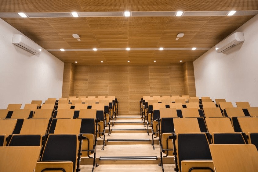 Sala konferencyjna z drewnianą boazerią, rzędami krzeseł i klimatyzacją. Oświetlenie sufitowe. Widok na salę z perspektywy widza.