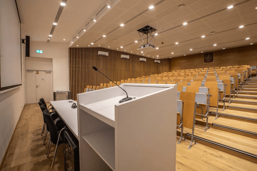 Nowoczesna aula uniwersytecka z drewnianymi elementami, oświetleniem LED i białą mównicą z mikrofonem. Widoczne rzędy krzeseł i godło Polski na ścianie.