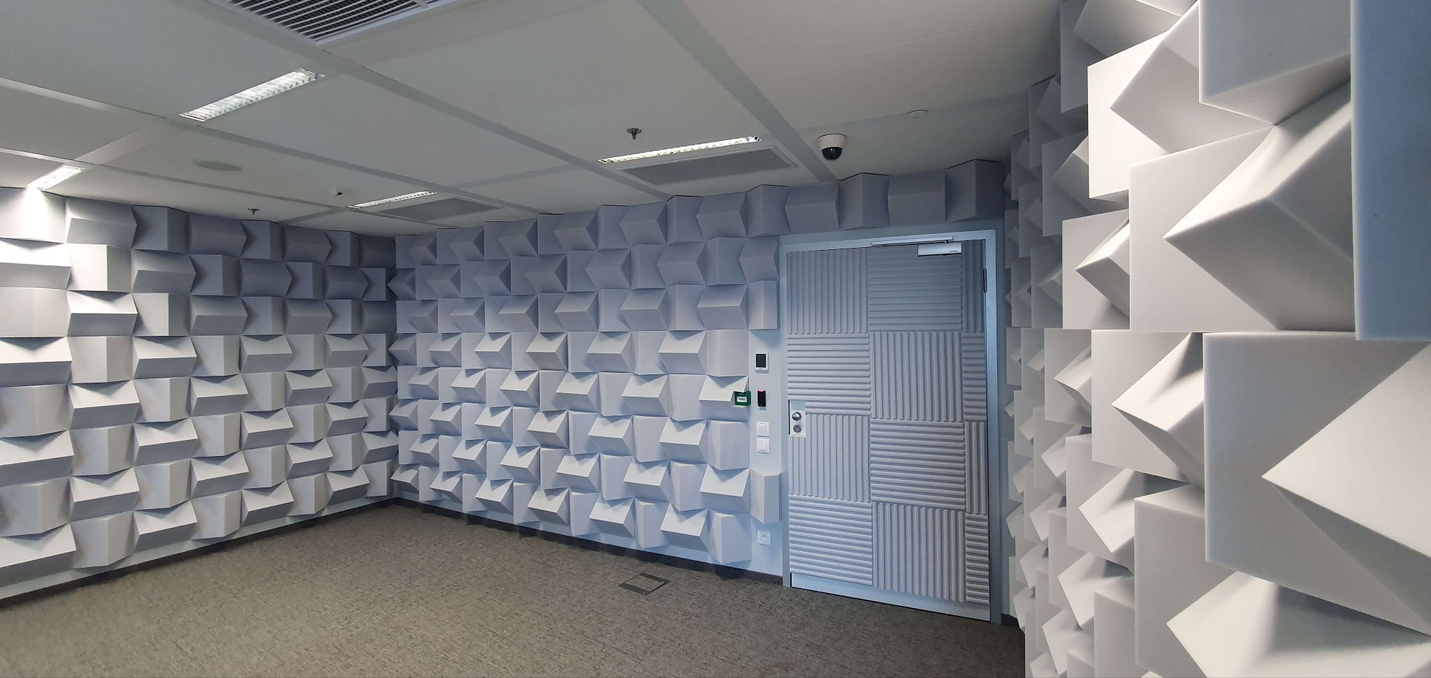 Pokój z panelami akustycznymi 3D na ścianach i suficie, szara wykładzina podłogowa, drzwi z poziomymi żłobieniami. Oświetlenie sufitowe.