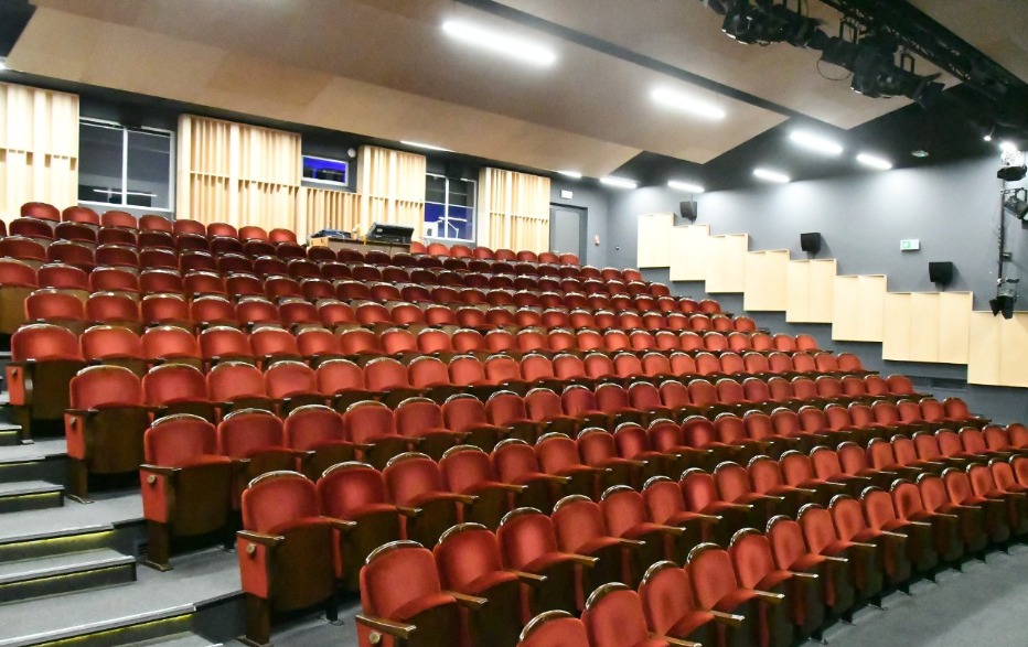 Sala widowiskowa z rzędami czerwonych foteli, oświetleniem scenicznym i panelami akustycznymi na ścianach. Widok na puste audytorium przed wydarzeniem.