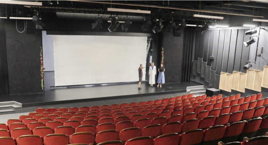 Widok na scenę teatru z rzędami czerwonych foteli. Na scenie trzy osoby, oświetlenie punktowe. Ściany w ciemnych kolorach, akustyczne panele po prawej.