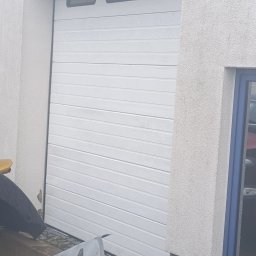 Adam Łucyk - Biała brama garażowa segmentowa z trzema oknami, osadzona w jasnej elewacji o fakturze baranka. Widoczne ślady zabrudzeń u dołu bramy i na elewacji.