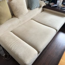 bluestart.kak - Jasna, narożna kanapa z plamami na siedzisku, widoczna z boku. Na kanapie poduszki w różnych kolorach. Ciemna podłoga z paneli.