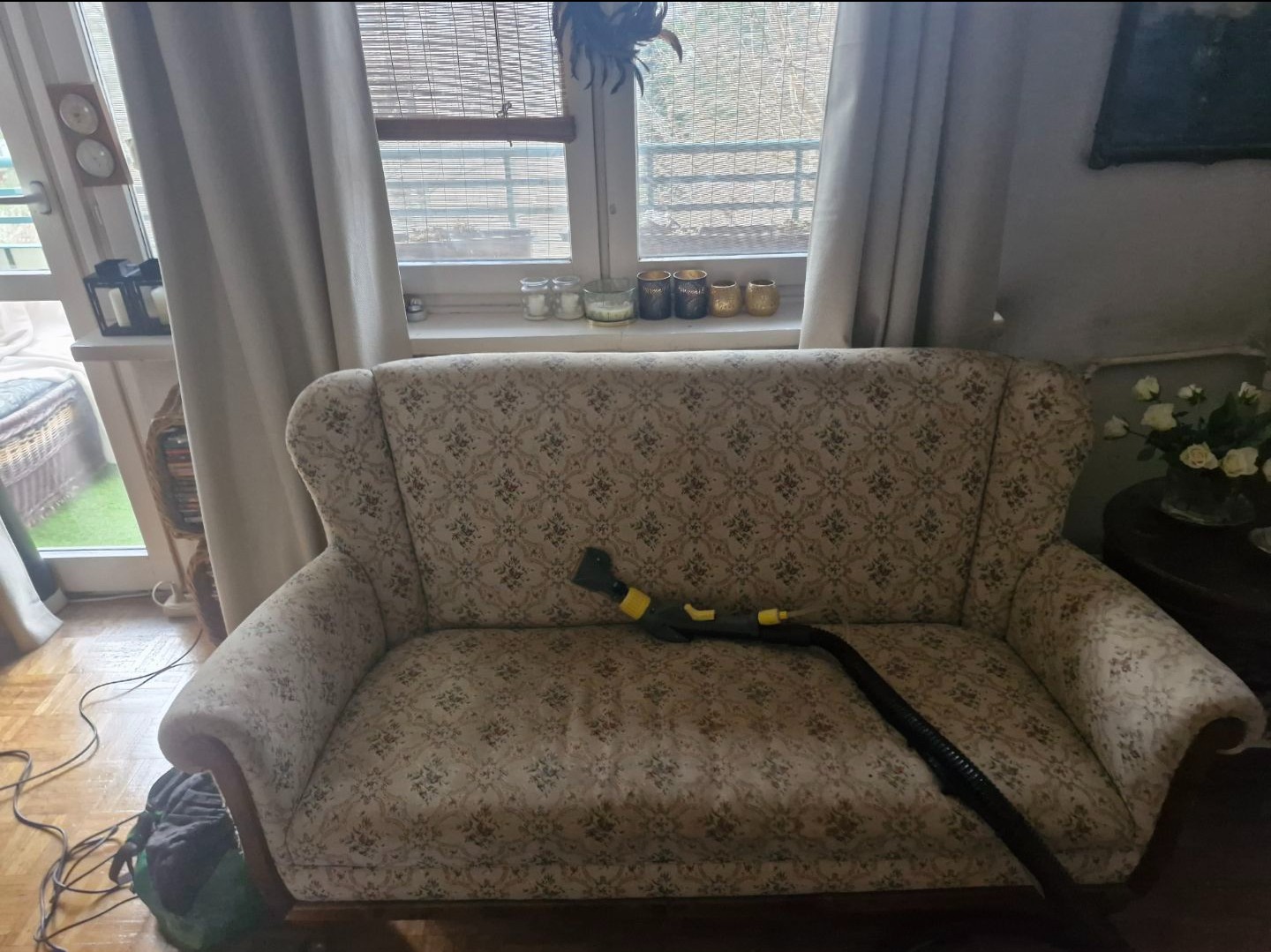 Wnętrze z sofą w kwiaty czyszczoną parowo. Dysza parowa na siedzisku, okno w tle. Widoczne ślady czyszczenia.