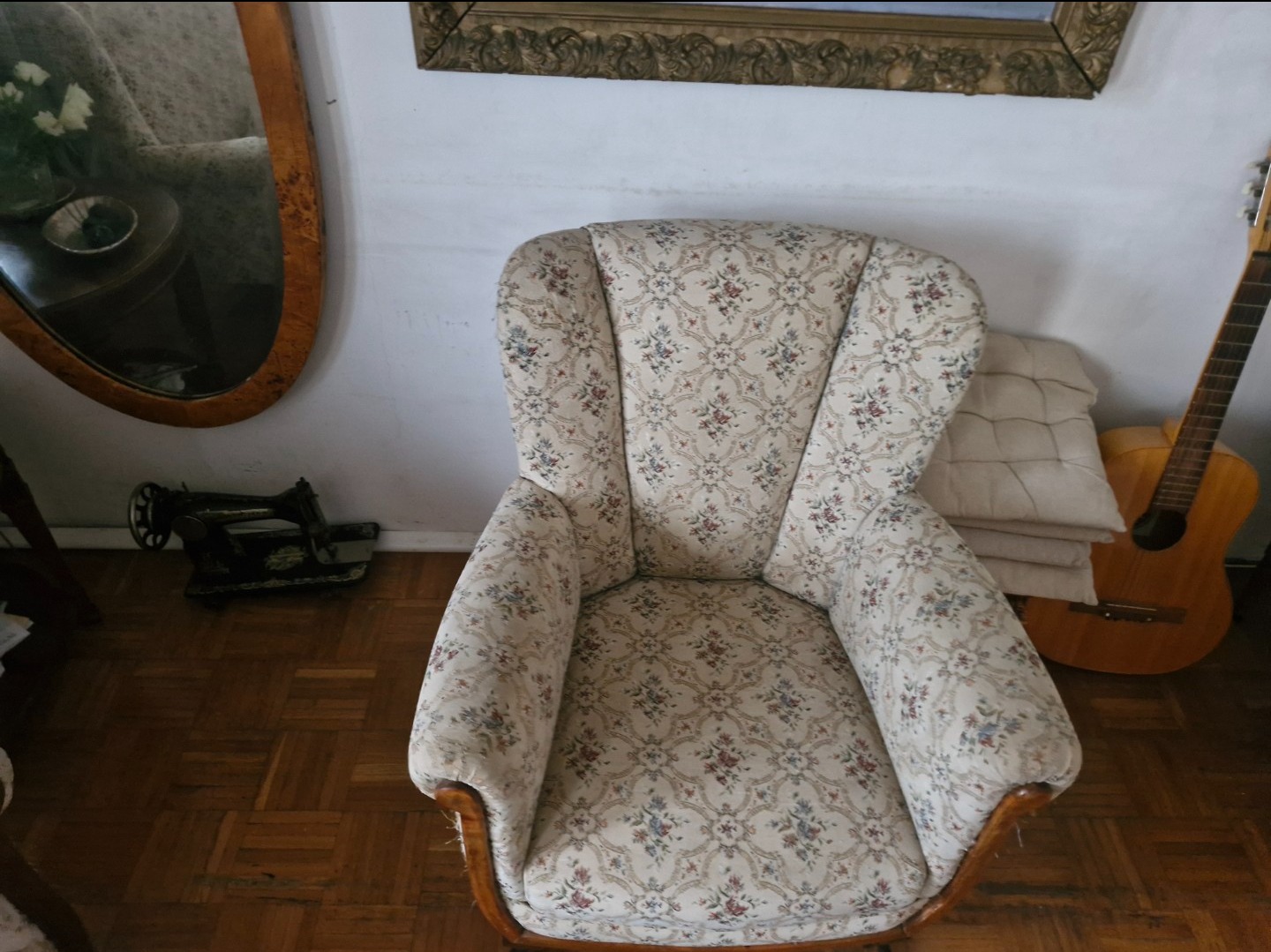 Wysokie krzesło tapicerowane w kwiaty, z drewnianymi nogami, stoi na parkiecie. W tle lustro w złotej ramie, gitara i stara maszyna do szycia.
