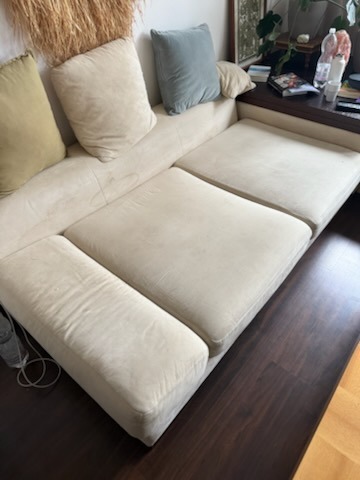 Jasna, narożna kanapa z plamami na siedzisku, widoczna z boku. Na kanapie poduszki w różnych kolorach. Ciemna podłoga z paneli.