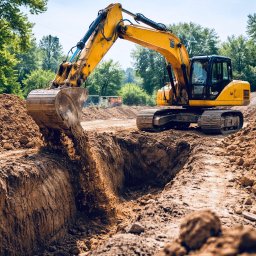 Royal Garden - Żółta koparka w trakcie wykopu głębokiego rowu w ziemi, z widocznymi śladami gąsienic i stertami ziemi po bokach. W tle zieleń drzew i błękitne niebo.