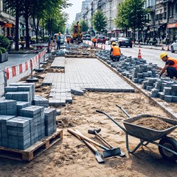 Royal Garden - Układanie szarej kostki brukowej na ulicy w mieście. Widoczni pracownicy w kamizelkach i kaskach, narzędzia pracy, paleta z kostką i częściowo ułożona nawierzchnia.