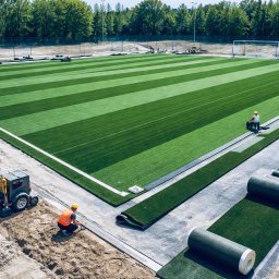 Royal Garden - Montaż trawy z rolki na boisku sportowym. Widoczne rolki trawy, pracownicy w kaskach i kamizelkach oraz maszyna do transportu materiałów. Częściowo ułożona murawa.