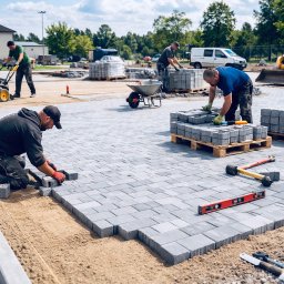 Royal Garden - Układanie szarej kostki brukowej na piasku. W tle pracownicy, ubijarka, palety z kostką, koparka i budynek. Praca wre na dużym placu.