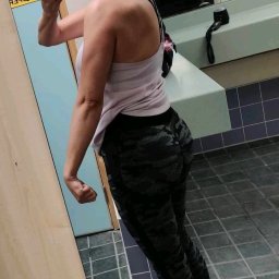 Ewa Iwańska - Kobieta w sportowym stroju pozuje w szatni, prezentując sylwetkę. Widoczny telefon w ręce, słuchawki bezprzewodowe i lustro. Ujęcie z boku, podkreślające muskulaturę ramienia.