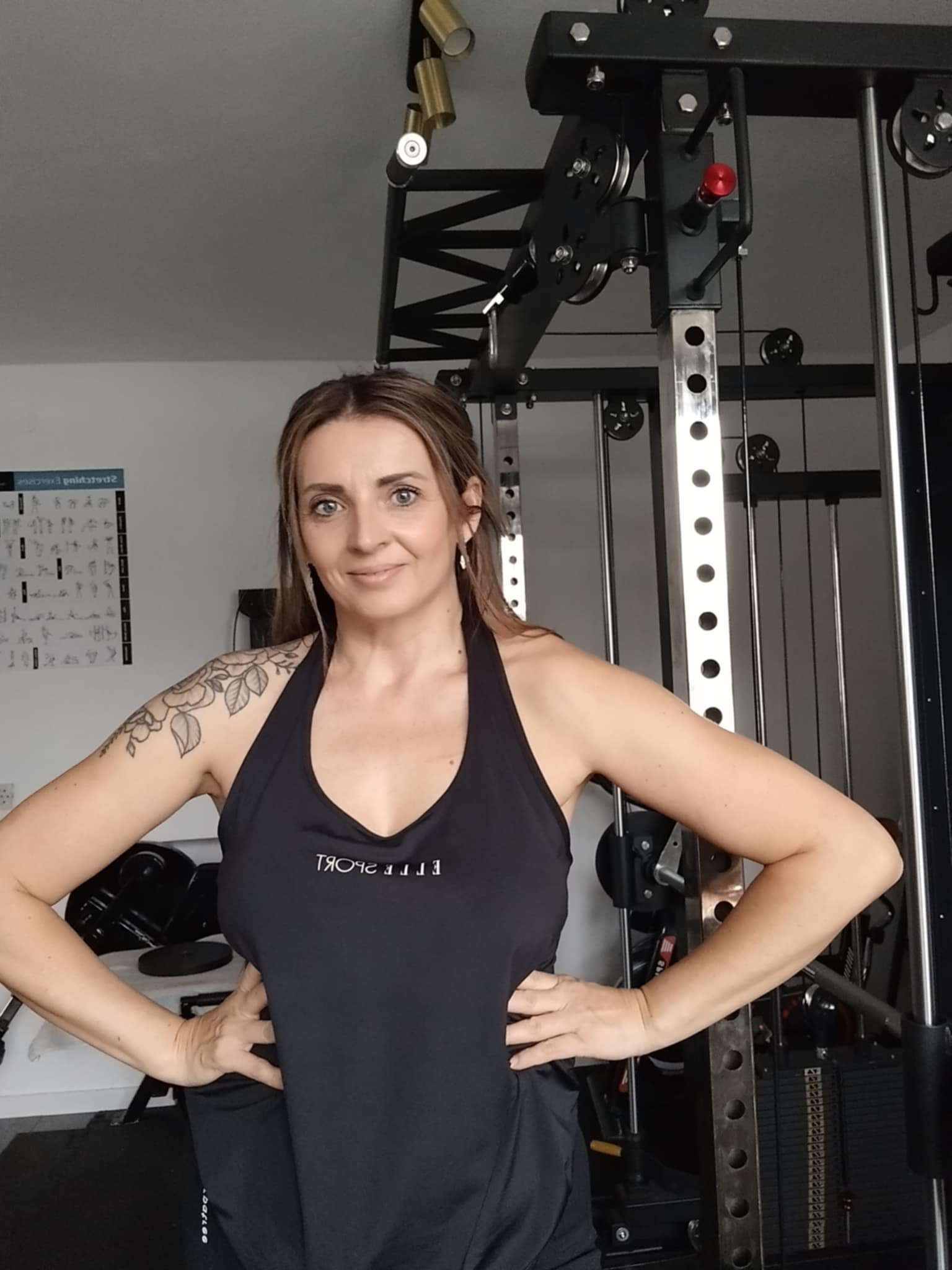 Kobieta z tatuażem na ramieniu, w czarnym stroju sportowym, pozuje na tle domowej siłowni z maszyną do ćwiczeń i plakatem z ćwiczeniami na ścianie.