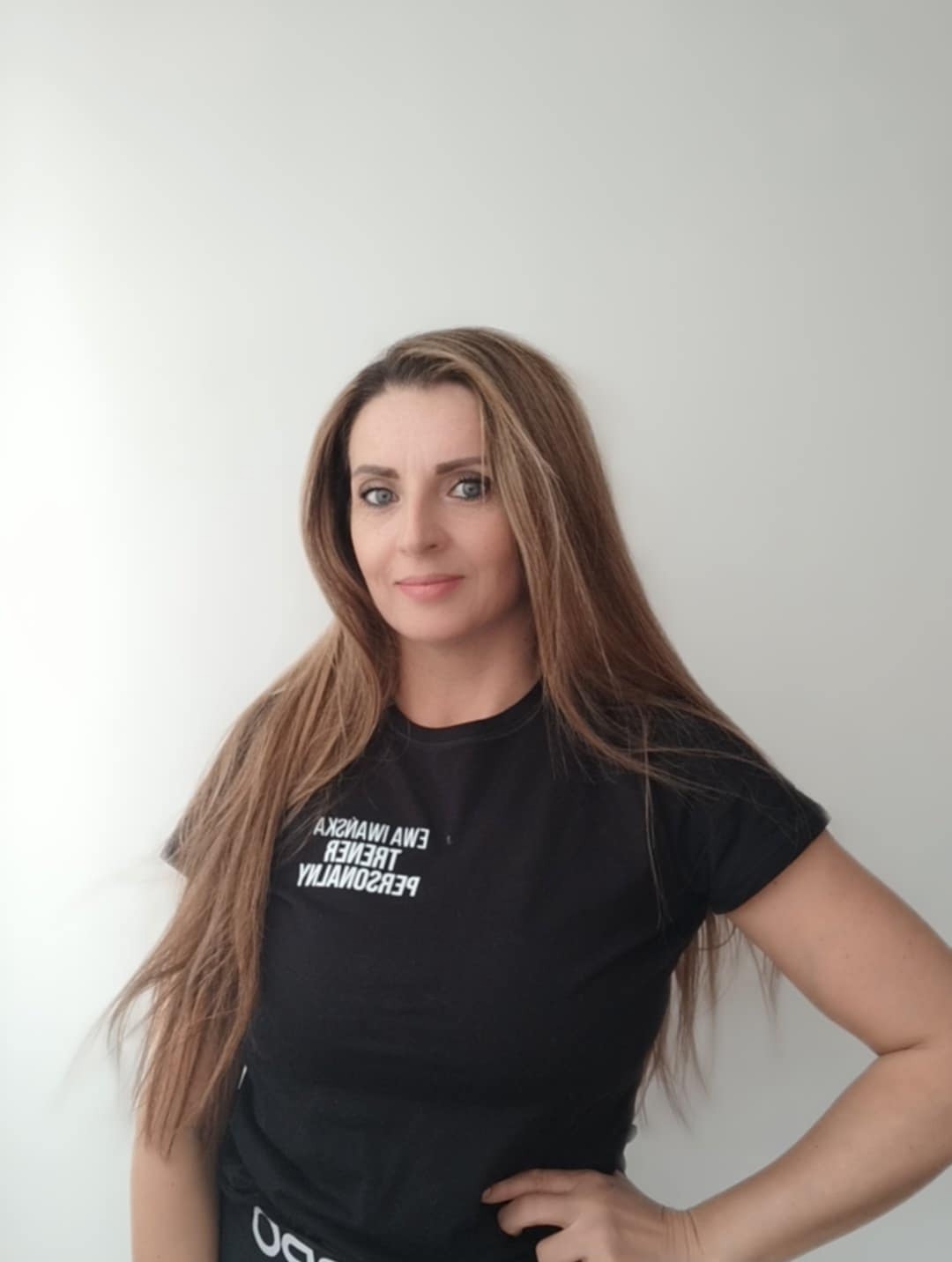 Portret kobiety z długimi włosami w czarnej koszulce z napisem 'Trener Personalny' w lustrzanym odbiciu, pozującej na jasnym tle.