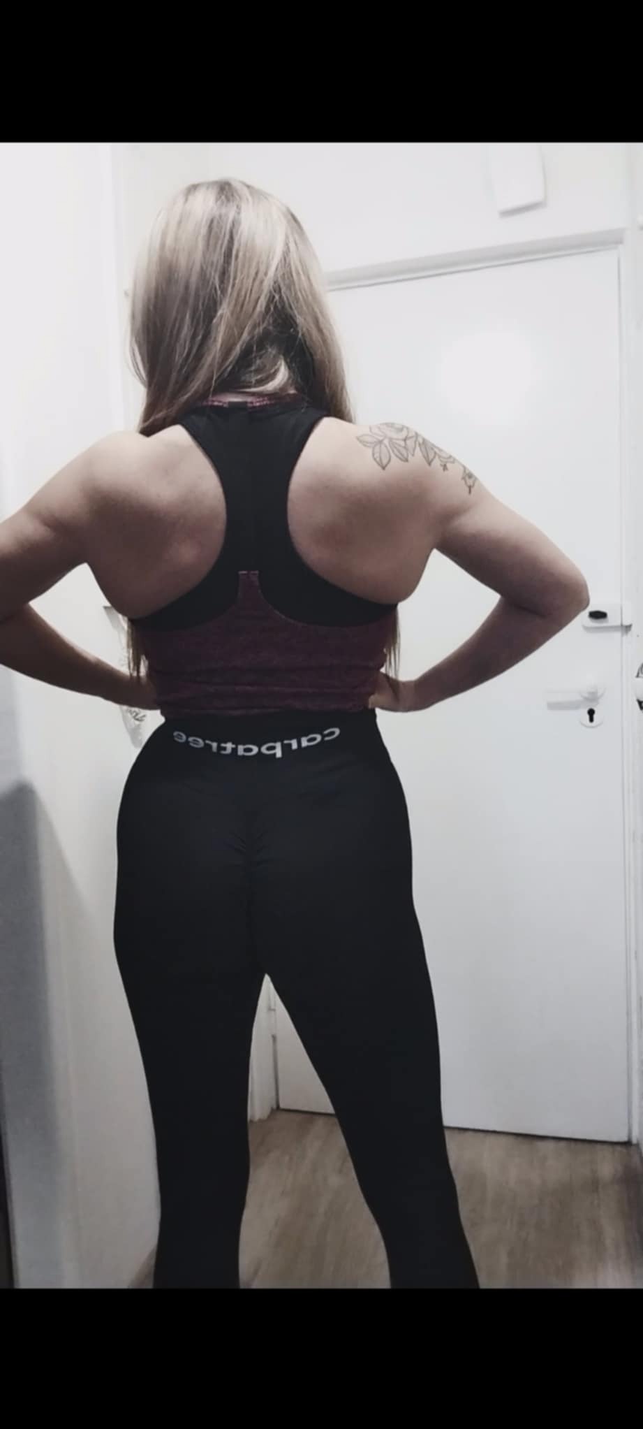 Kobieta z tatuażem na ramieniu, prezentująca muskulaturę pleców i ramion w sportowym stroju. Widok z tyłu, na jasnym tle drzwi.