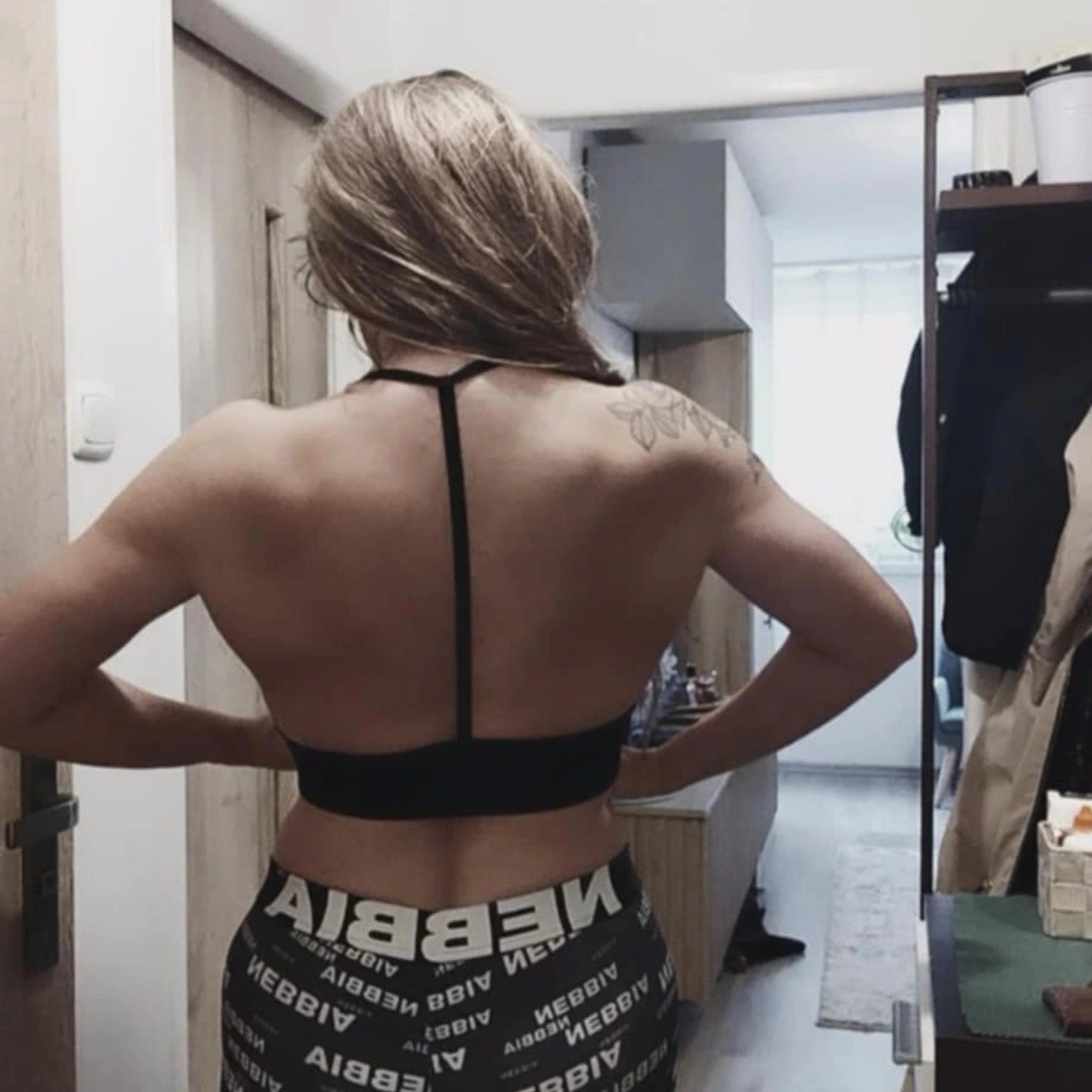 Kobieta w sportowym stroju, widok z tyłu, prezentująca umięśnione plecy i ramiona. Tatuaż na ramieniu. Ubrana w czarny top i legginsy z napisem.