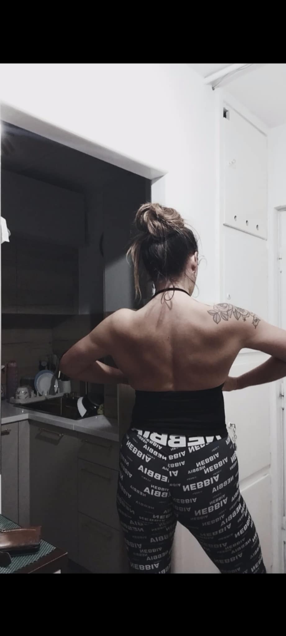 Kobieta tyłem, w sportowych legginsach z napisem, prezentuje umięśnione plecy i tatuaż na ramieniu w domowym otoczeniu. Widoczna kuchnia w tle.