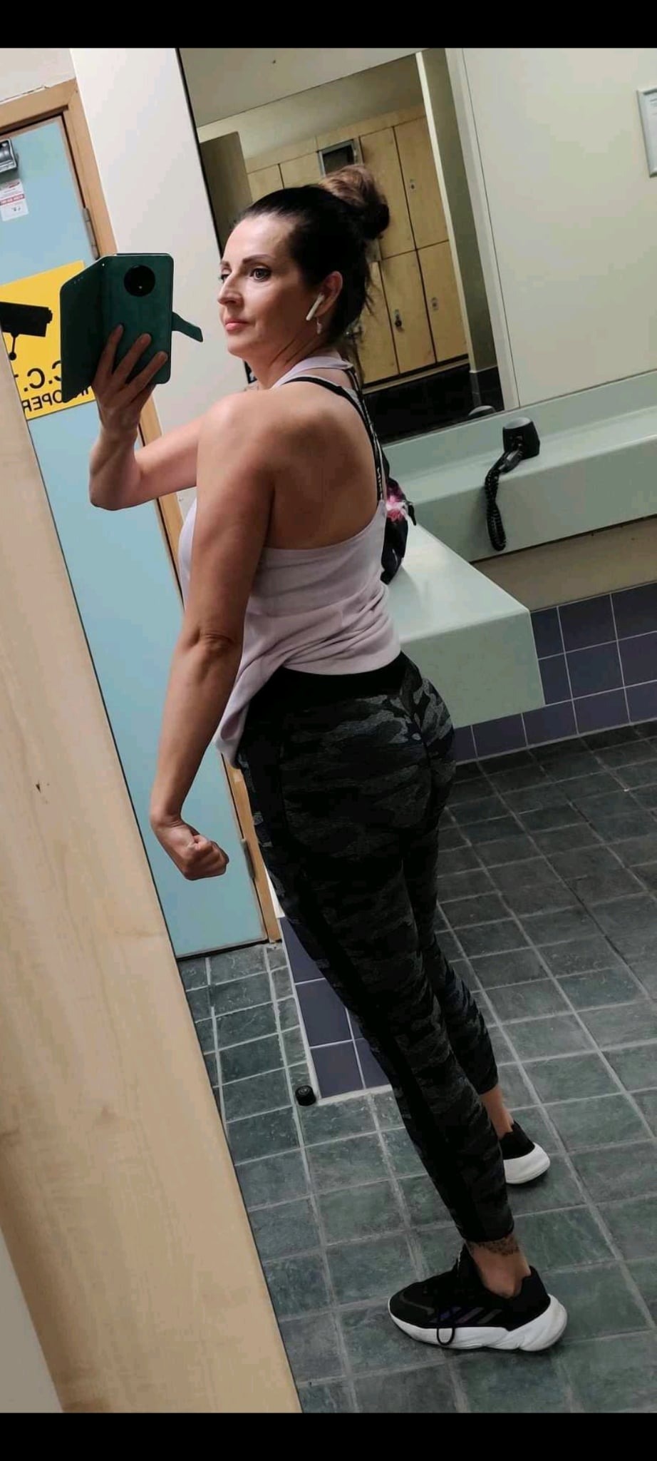 Kobieta w sportowym stroju pozuje w szatni, prezentując sylwetkę. Widoczny telefon w ręce, słuchawki bezprzewodowe i lustro. Ujęcie z boku, podkreślające muskulaturę ramienia.