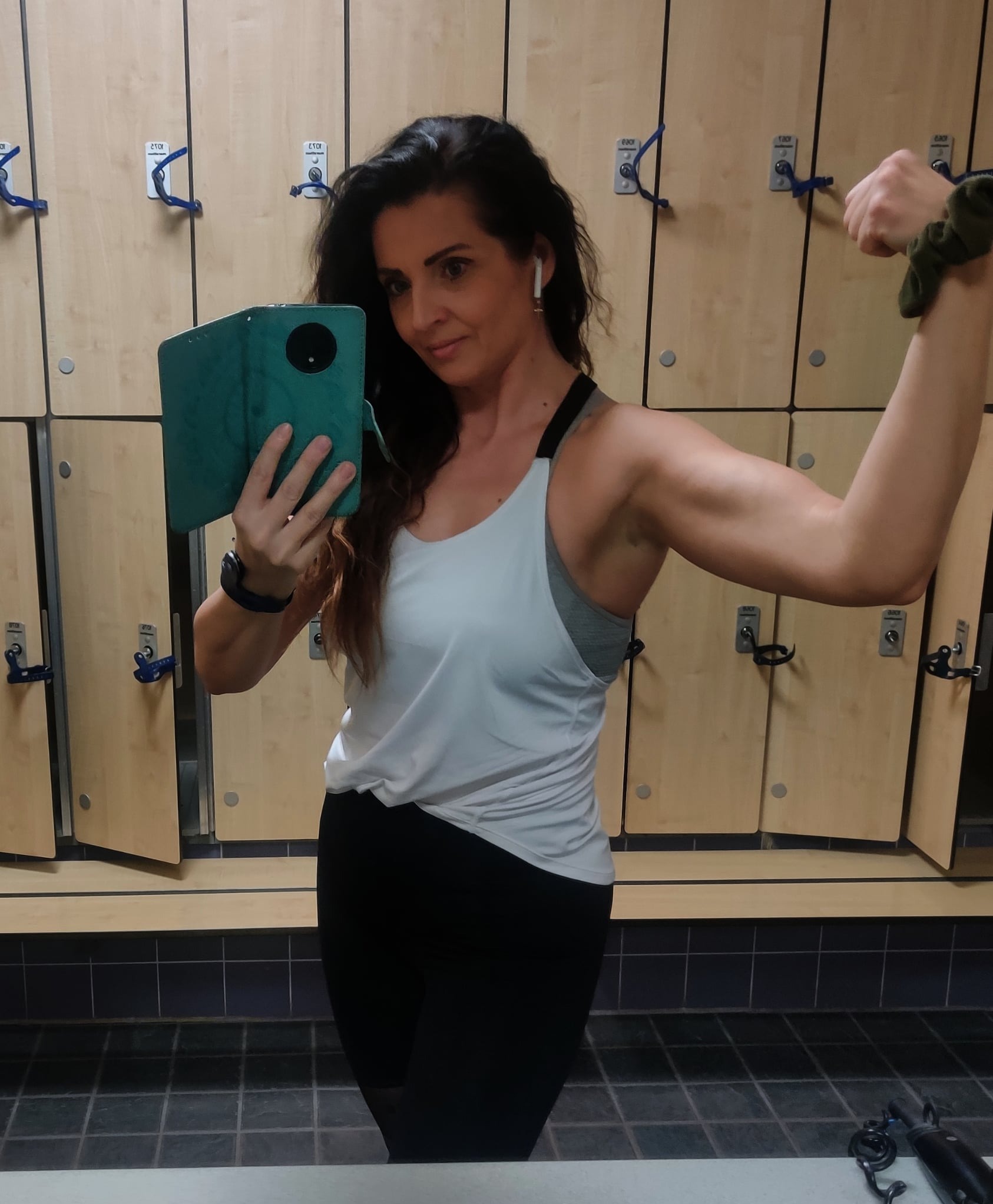 Kobieta w sportowym stroju robi selfie w szatni, prezentując napięty biceps. W tle rząd szafek z niebieskimi uchwytami i ciemna podłoga. Widoczny zegarek sportowy i słuchawki.