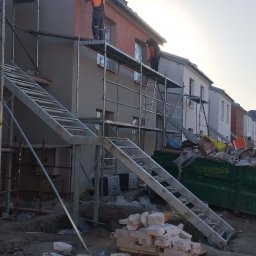 Vasyl Bihun - Prace elewacyjne w toku: robotnicy na rusztowaniu, fragment budynku z ociepleniem i tynkiem, gruz budowlany na ziemi. Widok na budowę w słoneczny dzień.