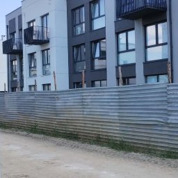 Vasyl Bihun - Nowoczesny budynek wielorodzinny z balkonami, wykończony w odcieniach szarości i bieli. Widok zza metalowego ogrodzenia placu budowy, w tle błękitne niebo.