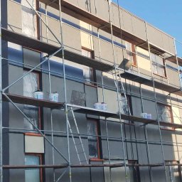 Vasyl Bihun - Budynek w trakcie prac elewacyjnych: szare ściany, rusztowania, farby. Okna zabezpieczone folią. U dołu hałda ziemi. Słoneczny dzień.