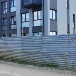 Vasyl Bihun - Nowoczesny budynek wielorodzinny z balkonem, wykończony w odcieniach szarości, widoczny zza metalowego ogrodzenia placu budowy w słoneczny dzień.