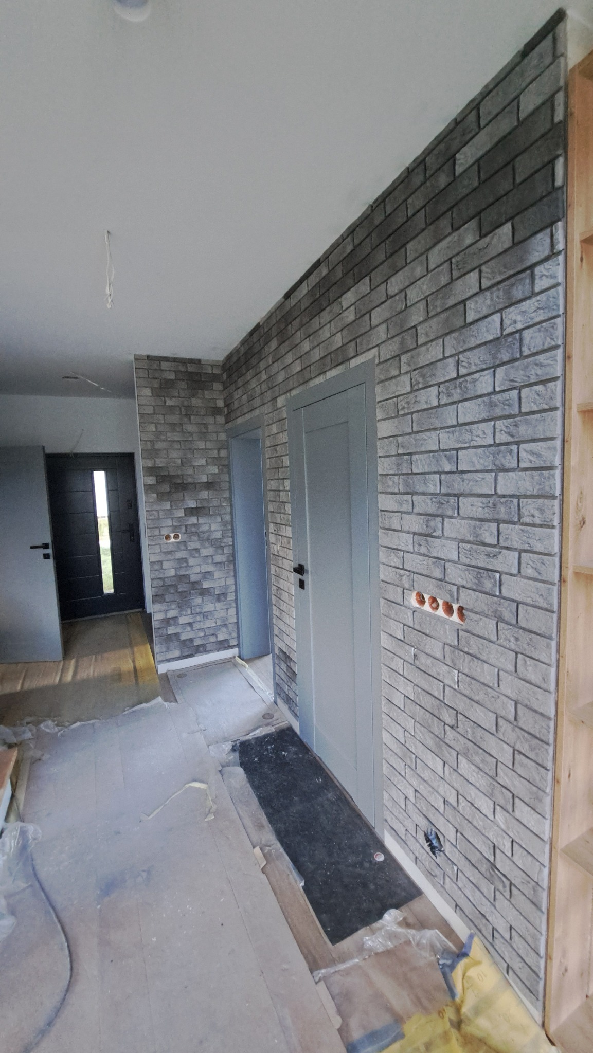 Korytarz po wykończeniu ściany imitacją cegły w odcieniach szarości. Widoczne szare drzwi i fragment drewnianego regału. Podłoga zabezpieczona folią malarską.