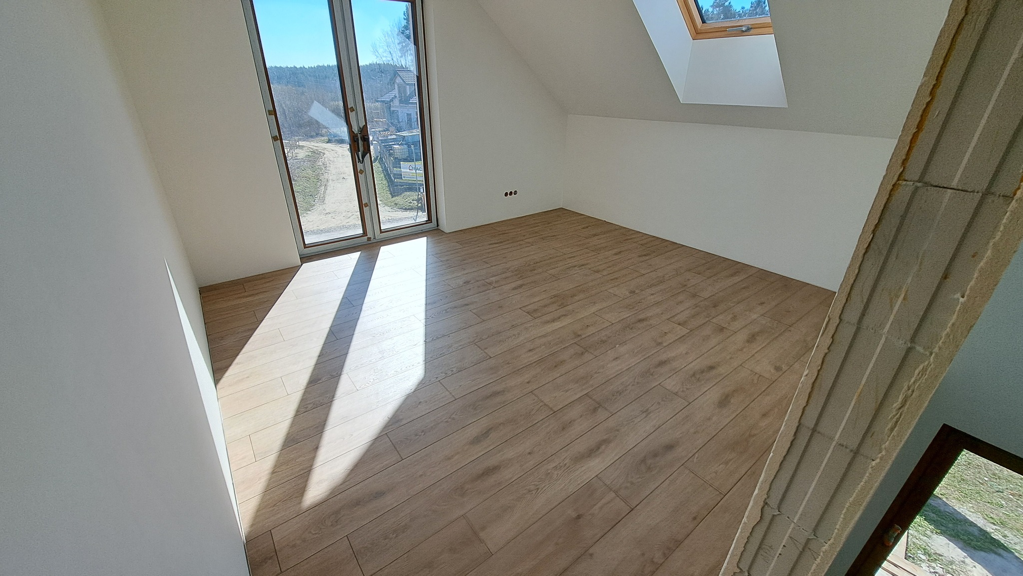 Jasne panele podłogowe w pokoju na poddaszu z oknem dachowym i dużym oknem balkonowym. Świeżo pomalowane ściany na biało. Widoczny fragment surowej ściany.
