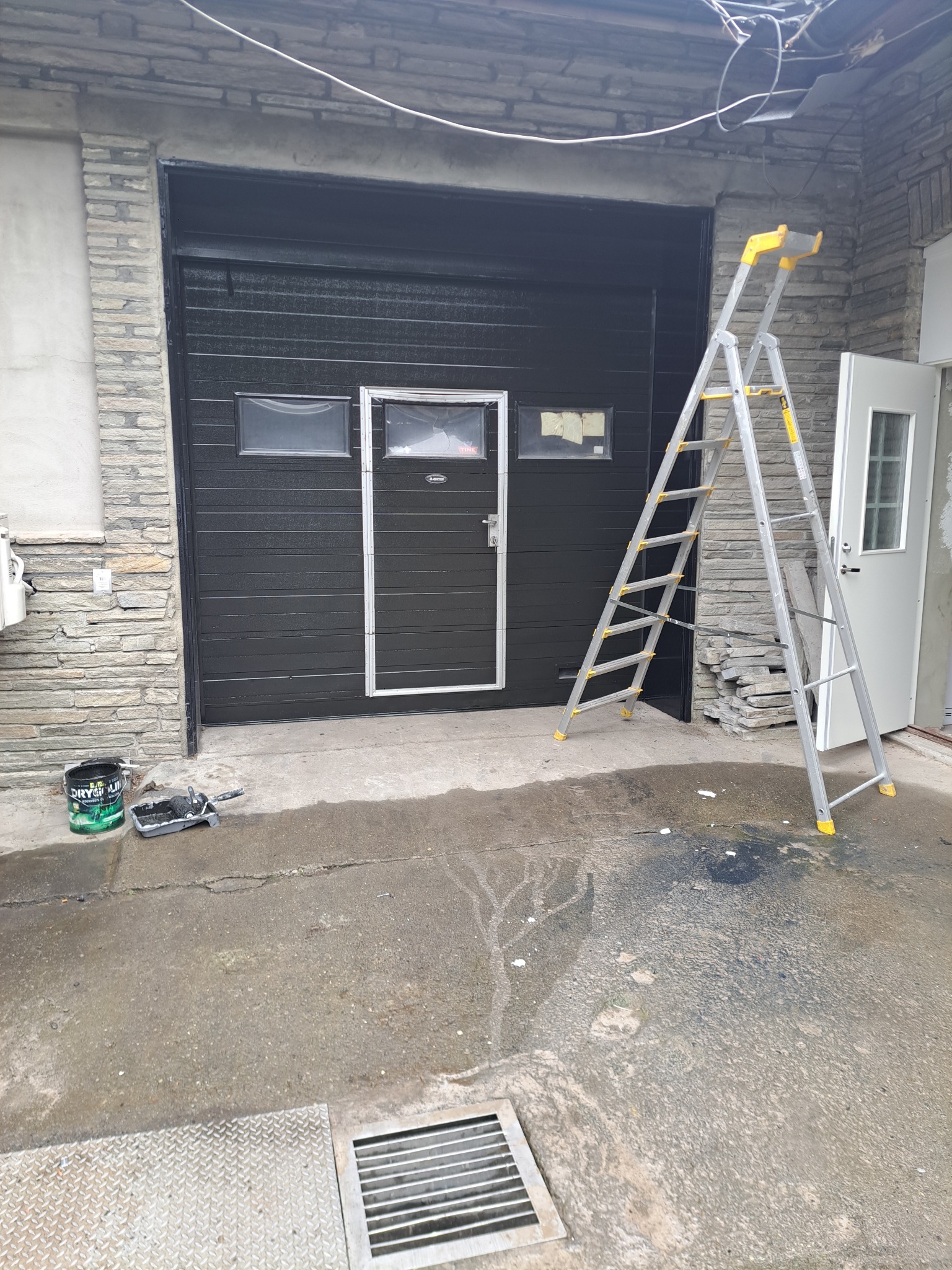 Świeżo pomalowana na czarno brama garażowa z oknami, drabina oparta o ścianę, puszka farby i wałek malarski obok. Kamienna elewacja budynku.