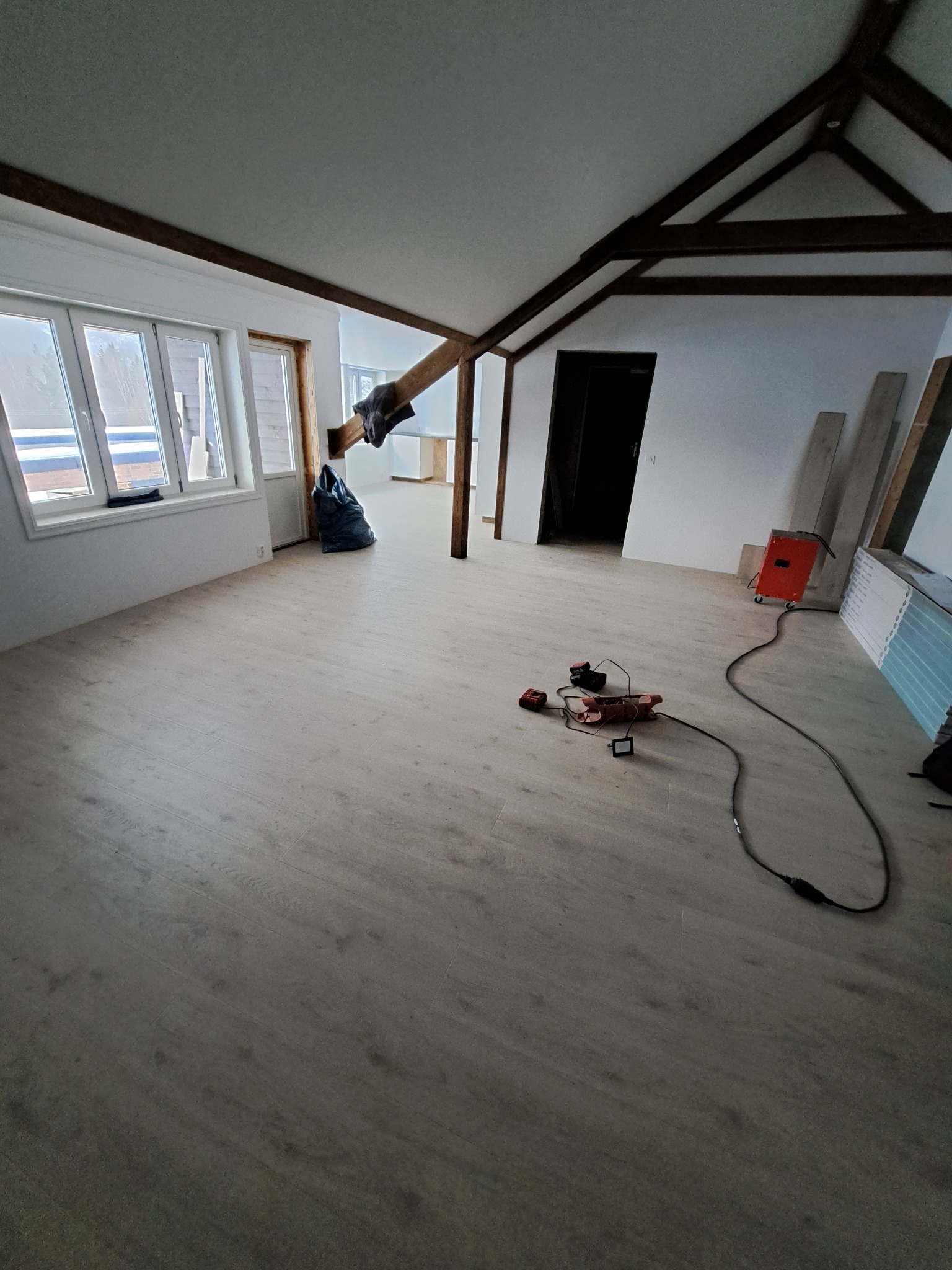 Jasna podłoga z paneli w pokoju ze skośnym sufitem i drewnianymi belkami. Widoczne narzędzia i materiały budowlane. W tle okno i drzwi.