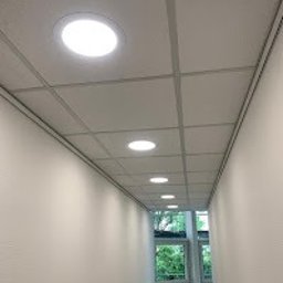Maciej Nowicki Instalacje Elektryczne - Równomiernie rozmieszczone, okrągłe lampy sufitowe LED w korytarzu z oknem. Białe ściany i sufit podwieszany z widoczną kratownicą. Ujęcie z dołu, perspektywa zbieżna.