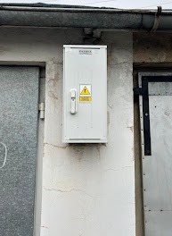 Biała skrzynka elektryczna zamontowana na szarej, lekko popękanej ścianie budynku. Widoczny symbol ostrzegawczy wysokiego napięcia i fragmenty metalowych drzwi po bokach.