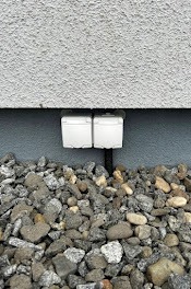 Dwa białe gniazdka elektryczne hermetyczne, zamontowane na elewacji budynku, z widocznym przewodem i kamiennym podłożem. Ujęcie z dołu, podkreślające detale instalacji.