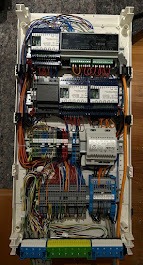 Rozdzielnica elektryczna z licznymi przewodami i modułami, widok z góry, ukazująca skomplikowaną instalację wewnątrz białej obudowy.