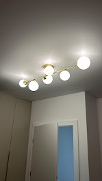 Nowoczesna lampa sufitowa z sześcioma kulistymi kloszami, zamontowana na złotej listwie, oświetlająca wnętrze z białymi ścianami i drzwiami.