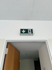 Podświetlony, zielony znak ewakuacyjny z piktogramem biegnącej osoby i strzałką w dół, zamontowany nad framugą drzwi w jasnym pomieszczeniu z podwieszanym sufitem.